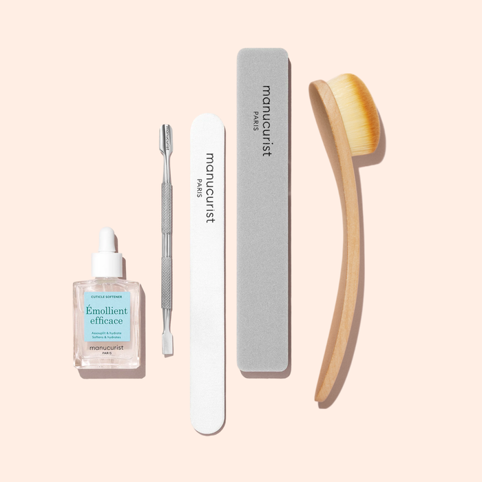 A set of manicure tools on a light background, featuring a cuticle softener, metal cuticle pusher, white nail file, grey buffer, and the Manucurist Dust Brush with vegan bristles.