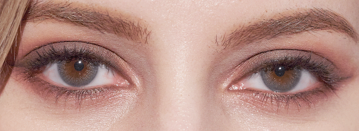 Close-up of a person's hazel eyes with subtle makeup, framed by natural brows. EyeCandys Attitude Glitter Grey contacts by EyeCandys enhance the eye color, while lighting highlights the skin texture around the eyes.