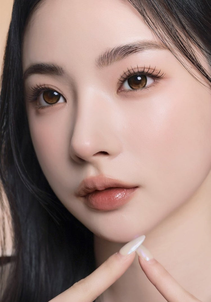 A female model with natural dark eyes wearing i-Sha Reborn Oak Haze contact lenses, complemented by subtle eyeshadow and peach lipstick