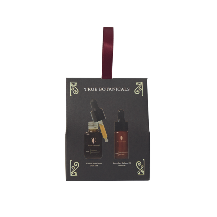 A dark gray, rectangular skincare gift box from True Botanicals styled as a hanging holiday ornament with a burgundy ribbon looped at the top. The front surface has minimal light-gold filigree accents in each corner and the brand name "TRUE BOTANICALS" printed in gold near the top. The center features realistic graphic prints of two items: an amber glass bottle of Chebula Active Serum on the left with its dropper exposed and dripping yellow-tinted liquid, and a bottle of Renew Pure Radiance Oil on the right