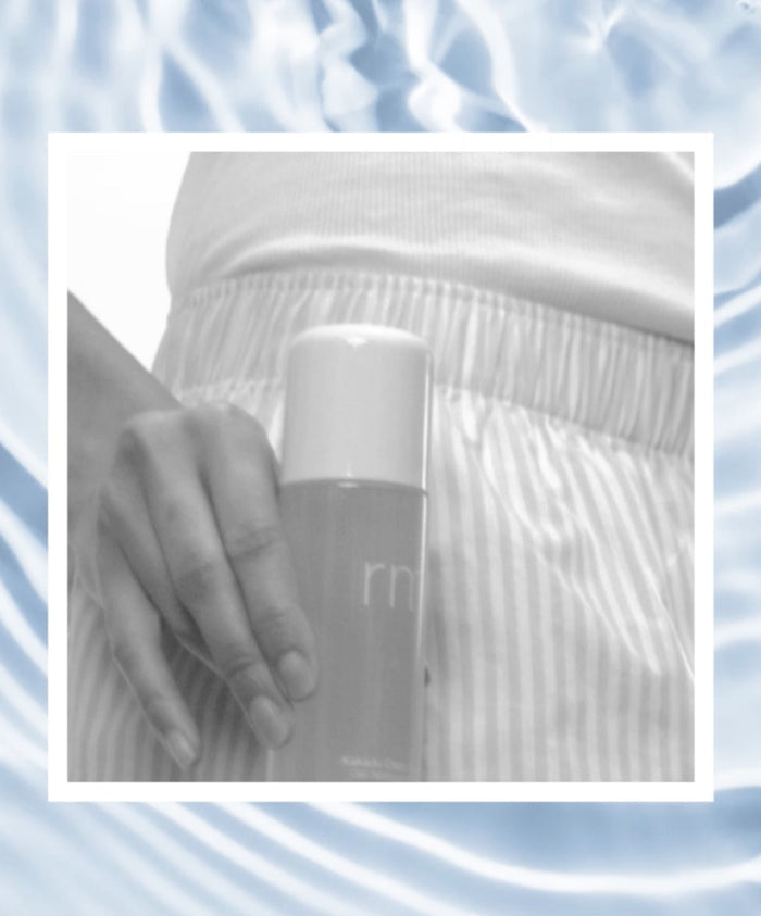 Close-up of a person wearing Striped Sleep Shorts holding a sleek skincare product bottle against a softly blurred blue water background, conveying calm and clean beauty.