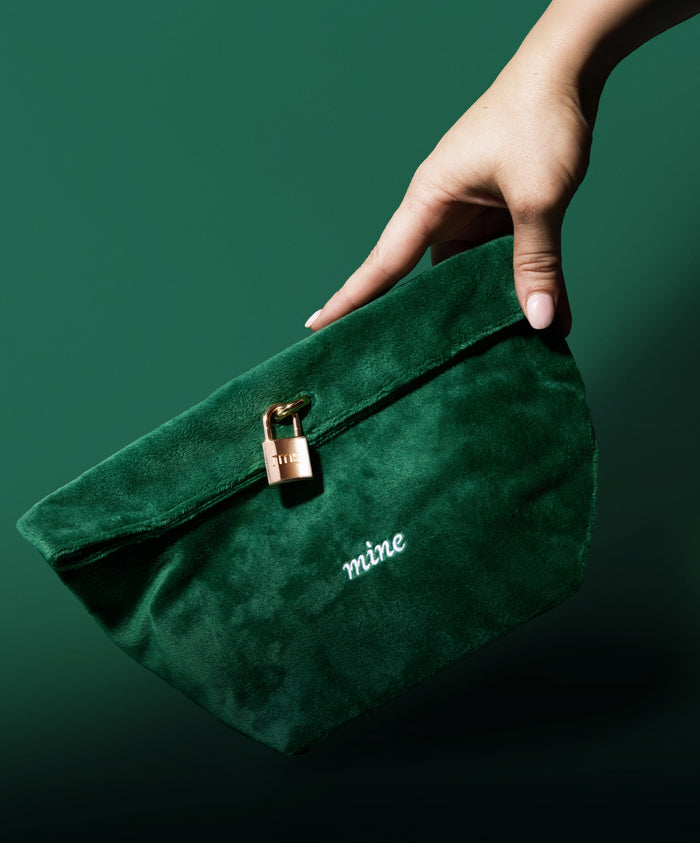 A hand holds The Emerald Luxe Pouch, a deep green velvet pouch with a gold clasp and the word "mine" embroidered on the front, against a matching green background.