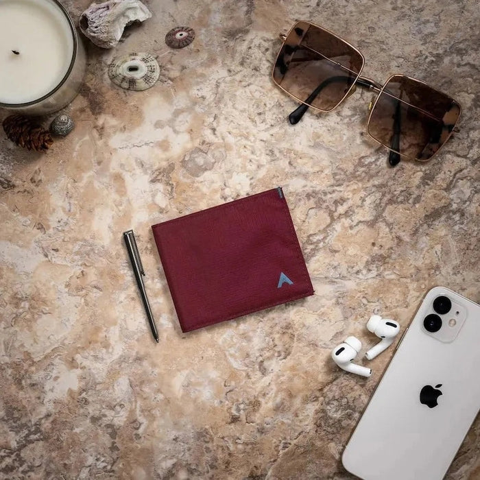 An ALLETT Sport Wallet in RFID-blocking mulberry nylon, along with a pen, sunglasses, candle, seashell, iPhone, and AirPods are neatly displayed on a marble surface.