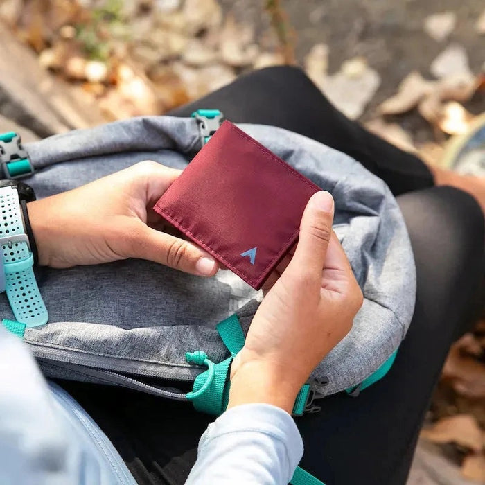 An ALLETT Sport Wallet in RFID-blocking mulberry nylon, along with a pen, sunglasses, candle, seashell, iPhone, and AirPods are neatly displayed on a marble surface.