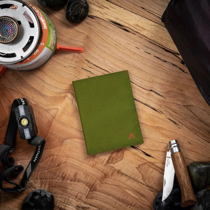 A Cala Green ALLETT RFID-blocking nylon wallet rests on a wooden surface beside a green notebook, portable stove, headlamp, black bag, pocket knife, camping gear, and round dark objects.