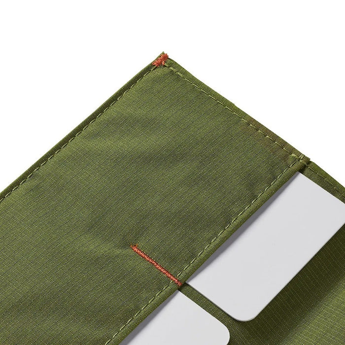 A Cala Green ALLETT RFID-blocking nylon wallet rests on a wooden surface beside a green notebook, portable stove, headlamp, black bag, pocket knife, camping gear, and round dark objects.