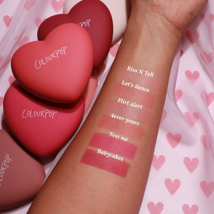 Babycakes pressed powder blush swatch anchors the bottom of a forearm lineup, delivering rich rosy mauve payoff with labeled shades above, as matte heart-shaped ColourPop compacts rest on pink heart fabric, highlighting the ColourPop Pressed Powder Blush.