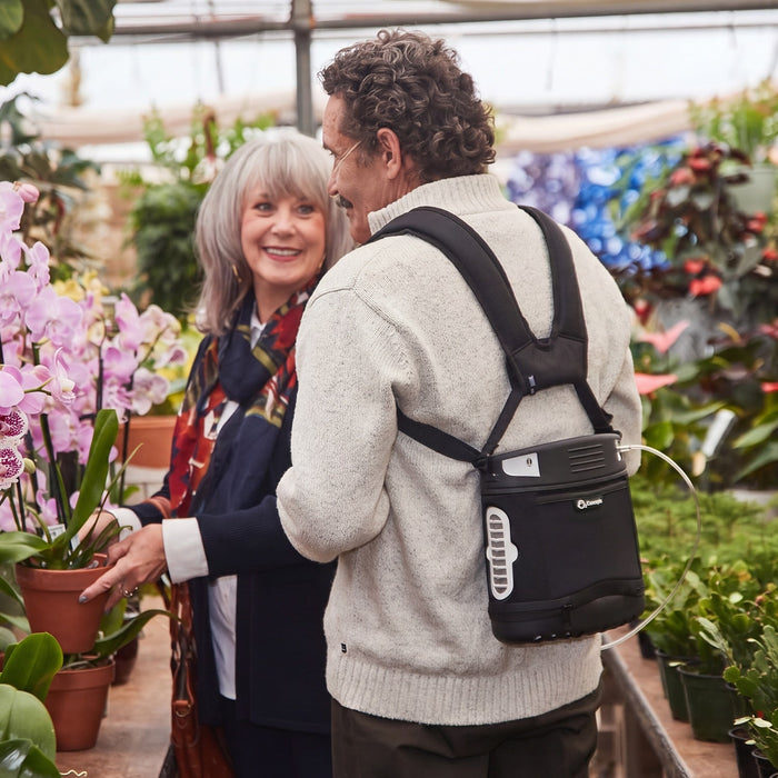 A man wearing a black backpack with an oxygen concentrator in it in a greenhouse, standing next to a woman who is looking at pink orchids.