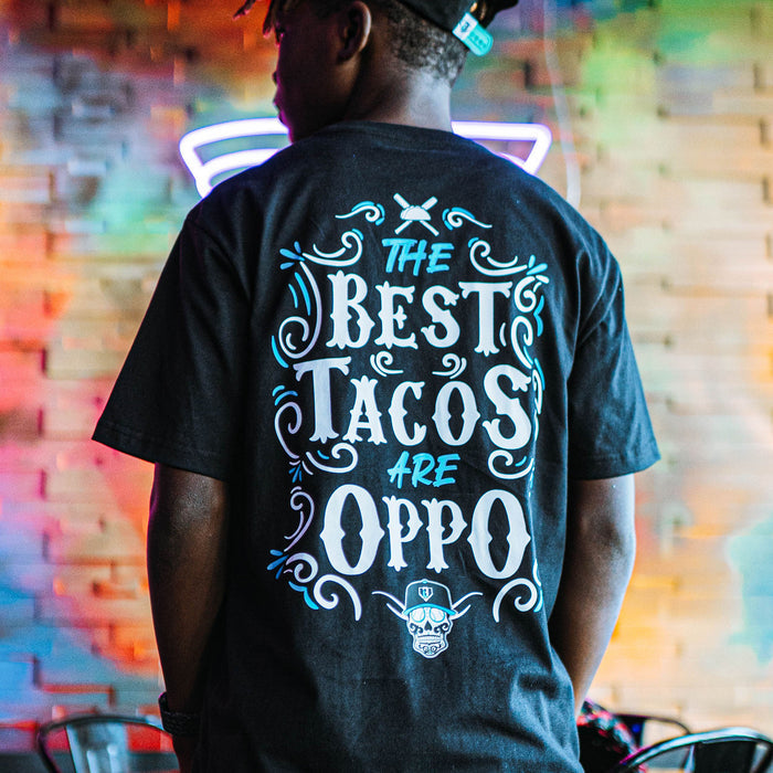 Person wearing a black t-shirt with 'The Best Tacos Are Oppo' text in a neon-lit setting
