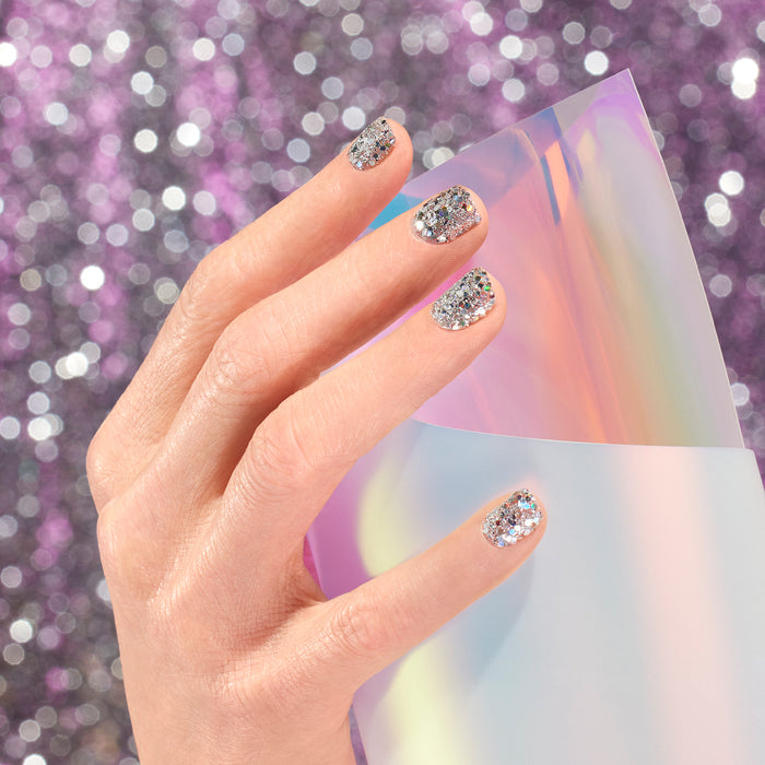 A hand with Manucurist Biodegradable Holo Glitter nail art holds a shiny, iridescent sheet against a background of sparkling purple bokeh lights.