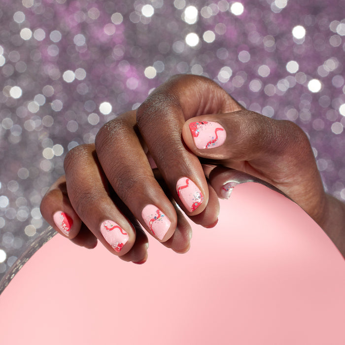 A hand with light pink nails, accented by red wavy lines and Manucurist’s Biodegradable Holo Glitter, holds a round pink mirror against a sparkly, blurred silver and purple background.
