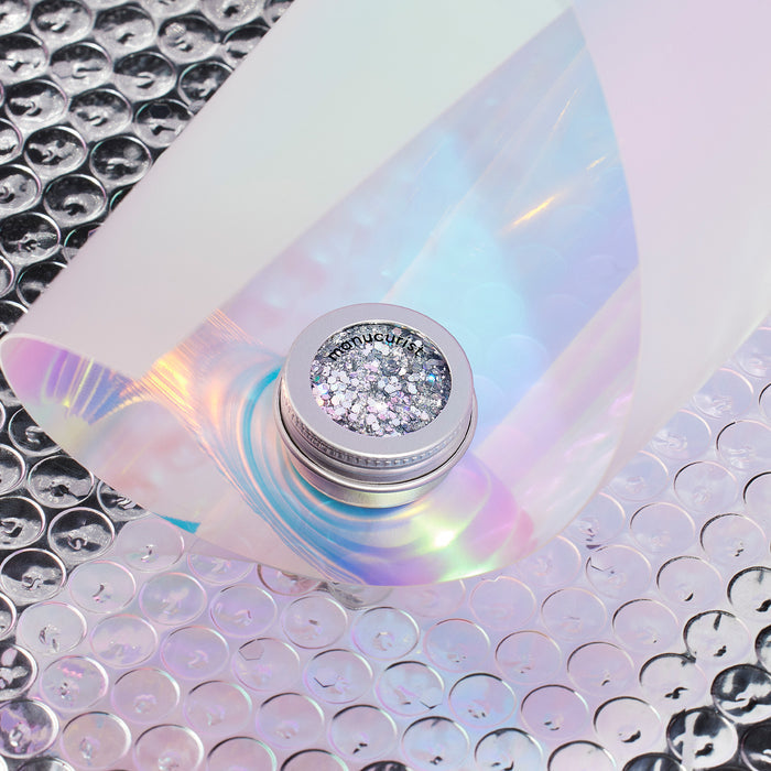 A small jar of Manucurist Biodegradable Holo Glitter rests on iridescent bubble wrap, partly covered by a translucent holographic sheet reflecting pastel hues—ideal for creating dazzling, eco-friendly nail art.