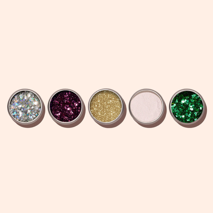 Five round pans of Manucurist Biodegradable Holo Glitter—silver, purple, gold, pale pink, and green—hold shimmering powders for creative nail art, neatly arranged on a light beige background.