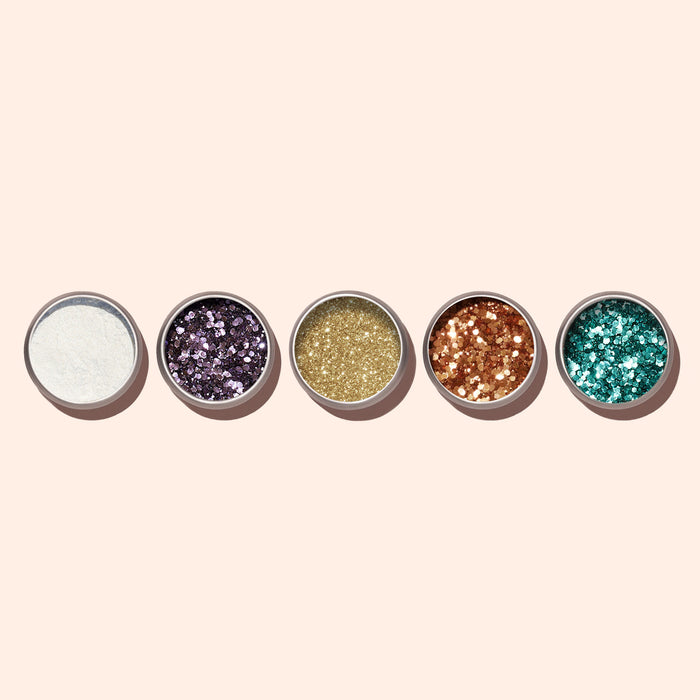 Five small tins of Manucurist Blue Glazed Powder—metal jars in white, purple, gold, copper, and pearly blue—are displayed in a row on a beige background. Perfect for adding sparkling shimmer to your manicure.