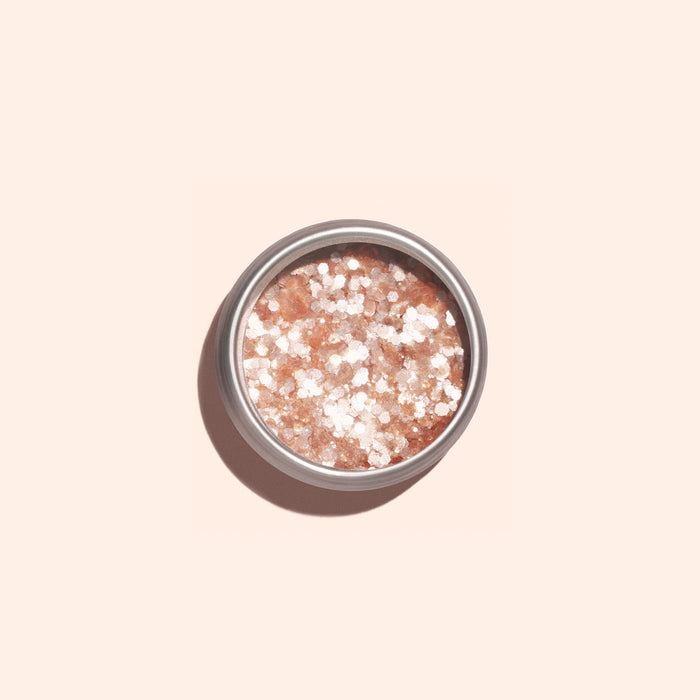 A round metal tin of Manucurist's Blush Biodegradable Glitter, featuring pink and white coarse bath salts with pale pink sparkle, sits on a light beige surface, casting a soft shadow to the left.