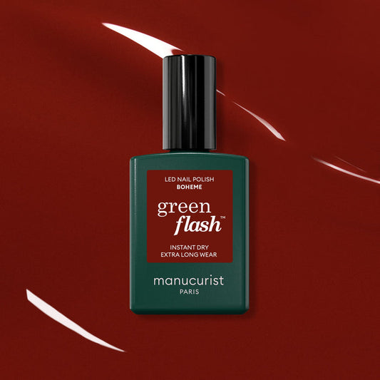 A bottle of Manucurist Bohème Green Flash LED polish—a Montmartre-inspired red-brown shade—rests on a glossy deep red surface with light reflections. The dark green bottle features a black cap and a red label.