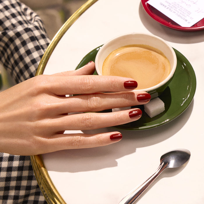 A hand with Manucurist's Bohème glossy red polish rests on a saucer holding an espresso cup, next to a spoon and sugar cubes, atop a marble table in Montmartre. The person wears black-and-white checkered pants.