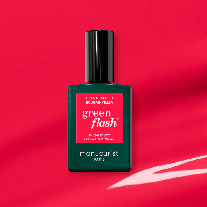 A bottle of Manucurist Bougainvillea Green Flash gel polish is shown on a red background with light reflections. The green bottle and red label highlight the pink undertone shade of this nail polish.