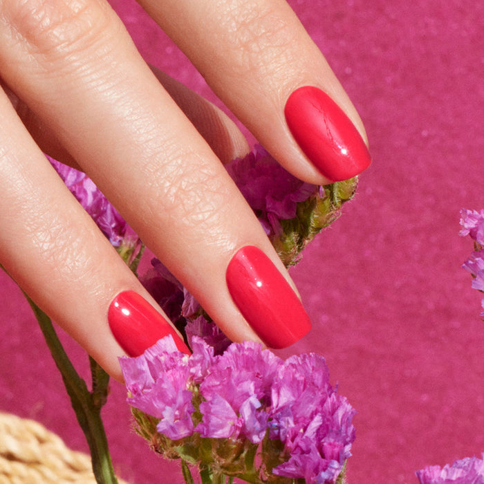 A hand with perfectly manicured nails painted in Manucurist's Bougainvillea gel polish holds a bunch of small purple flowers against a vibrant pink background.