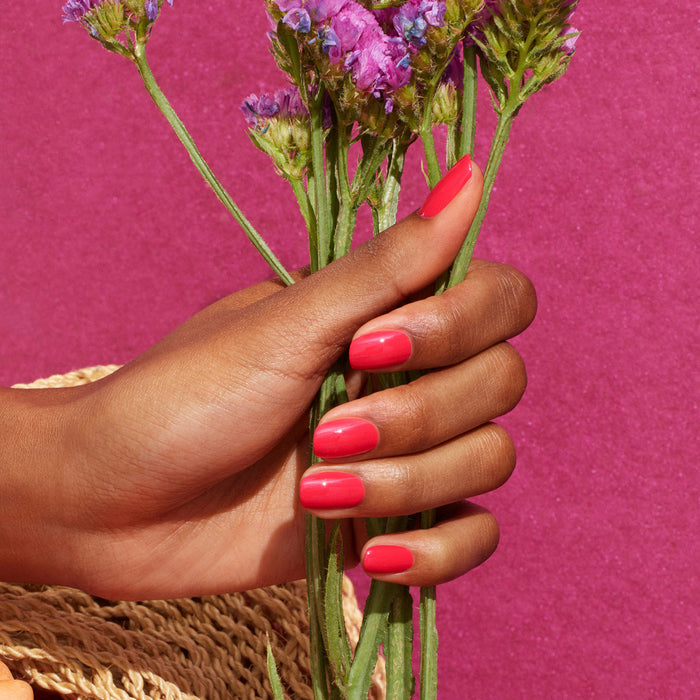 A hand with neatly manicured, glossy pink nails polished in Manucurist's Bougainvillea gel holds purple flowers against a vibrant pink background, with part of a woven hat visible in the corner.