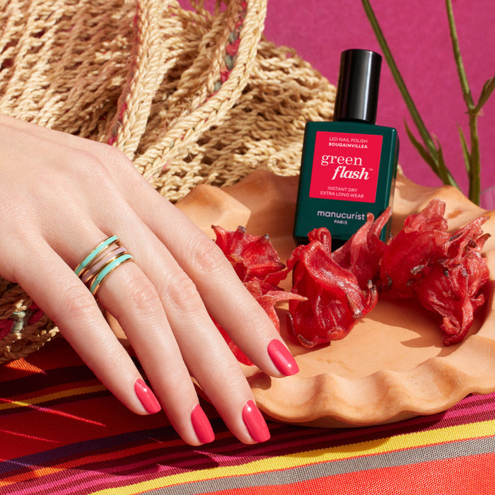 A hand with pink-painted nails in Manucurist’s Bougainvillea gel polish rests on a colorful striped surface, beside a green nail polish bottle, dried red flowers, and a woven straw bag. Three stacked gold rings adorn the fingers.