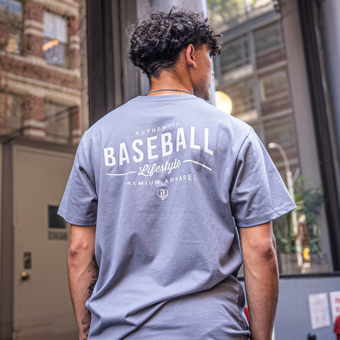 Person wearing a gray t-shirt with 'Authentic Baseball Lifestyle Premium Apparel' text on the back, standing in an urban setting.
