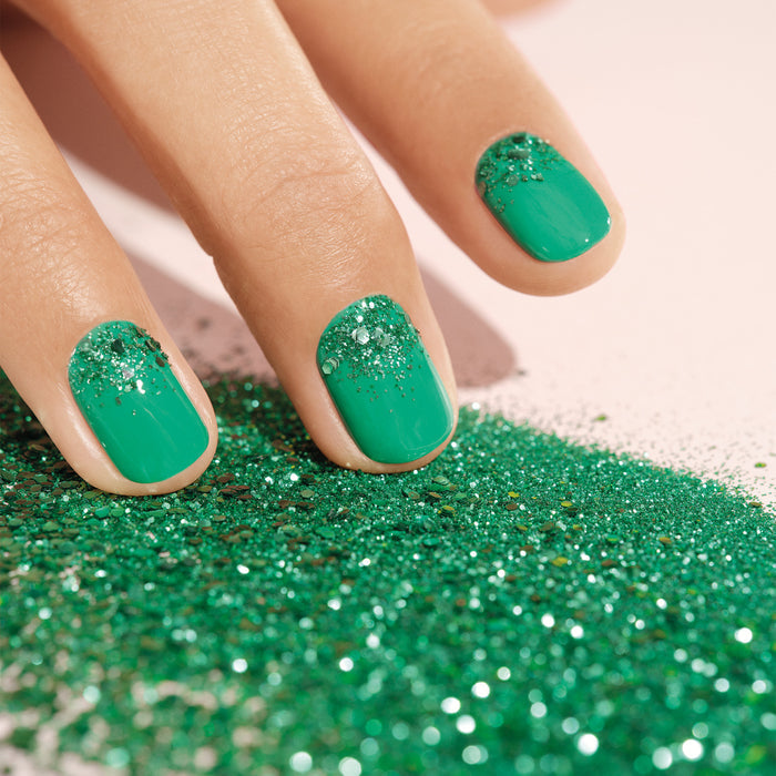 A hand with green nails, silver nail art, and Manucurist’s Cactus Biodegradable Glitter near the cuticles rests on a surface sprinkled with loose green glitter.