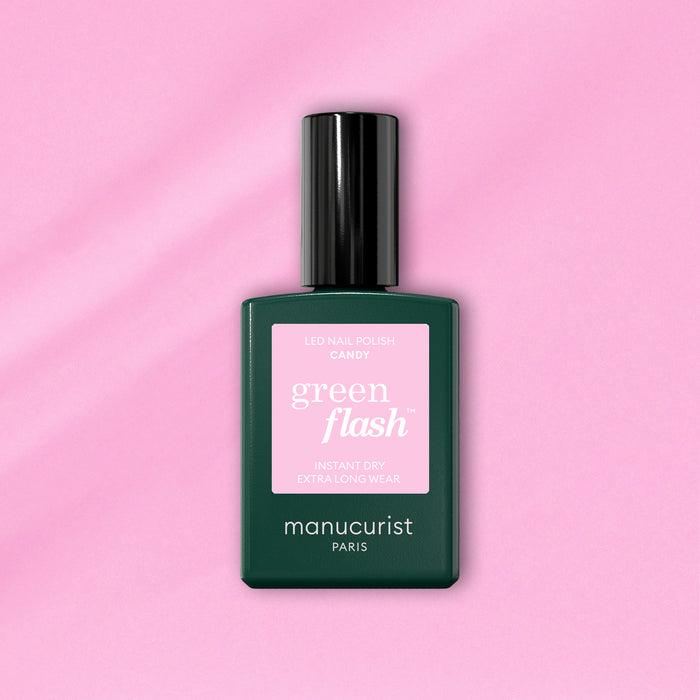 A bottle of Manucurist Candy Green Flash LED nail polish, with a pink label and dark green body, sits on a light pink background—ideal for those who love cool-toned LED manicures.