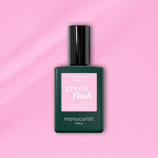 A bottle of Manucurist Candy Green Flash LED nail polish, with a pink label and dark green body, sits on a light pink background—ideal for those who love cool-toned LED manicures.