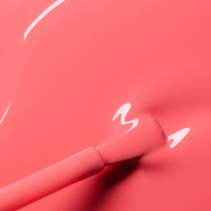 A close-up of a brush applying Manucurist's Capucine gel polish, a glossy coral pink shade with reflections of light shimmering on its smooth, wet surface.