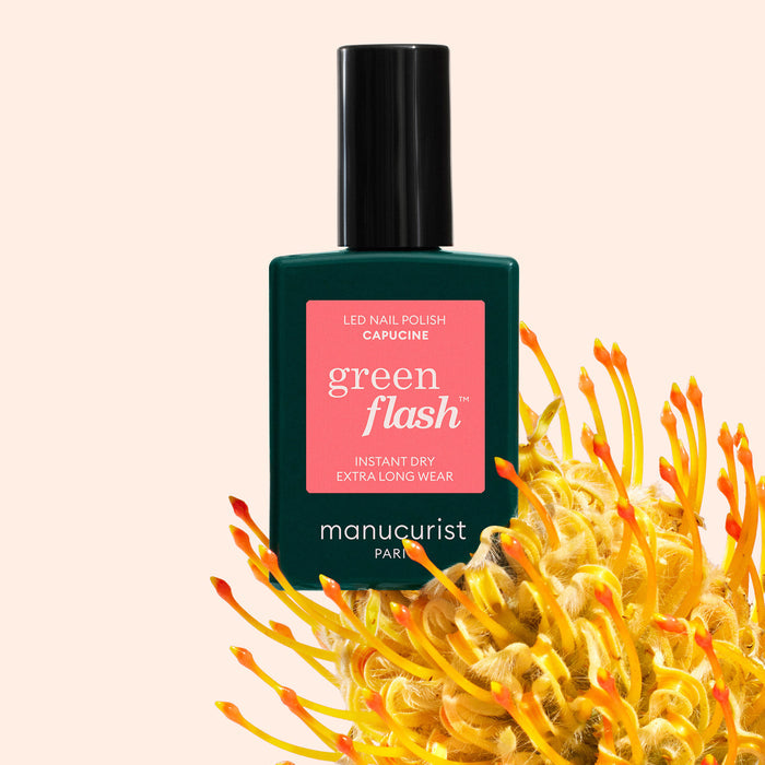 A bottle of Manucurist Capucine Green Flash gel polish sits among yellow-orange flowers on a light pink background. The bottle’s label is pink with white text.