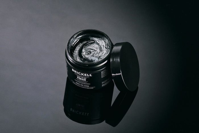 Black container of Brickell Men's pomade open with lid placed next to it on dark reflective surface.