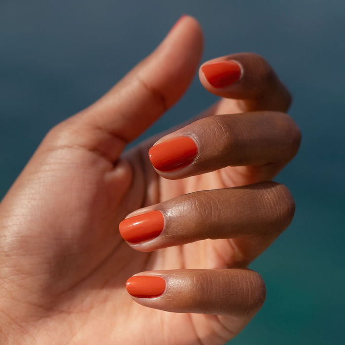 A close-up of a medium brown hand with neatly manicured nails painted in glossy orange Manucurist Color Refresher, which extends your manicure’s life, is shown against a blurred blue background.