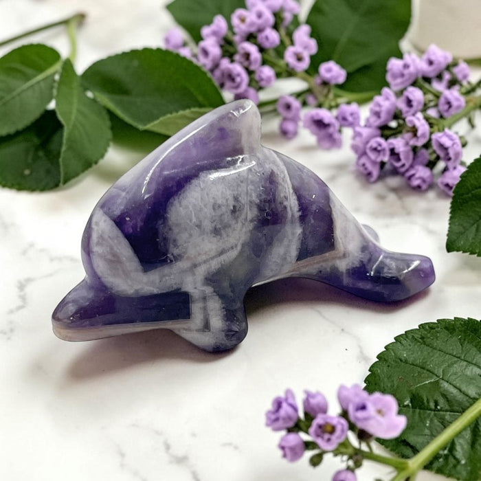 Purple and gray stone dolphin crystal on a marble surface with purple flowers and green leaves.