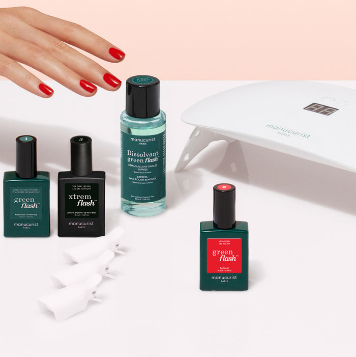 A hand with red nails is next to the Manucurist Discovery Kit, which includes nail polish, a vitaminized base coat, remover, a white LED lamp, and nail clips—everything needed for an ultra-glossy manicure.