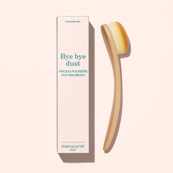 A Dust Brush by Manucurist with vegan bristles and a curved wooden handle sits beside its tall box labeled “Bye bye dust – Dust Brush” on a light beige background.