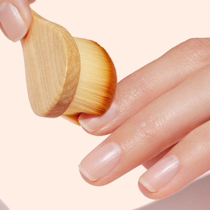 A close-up of a hand with clean nails being brushed by the Manucurist Dust Brush—featuring vegan bristles and a wooden handle—against a light peach background, perfect for eco-friendly manicure routines.