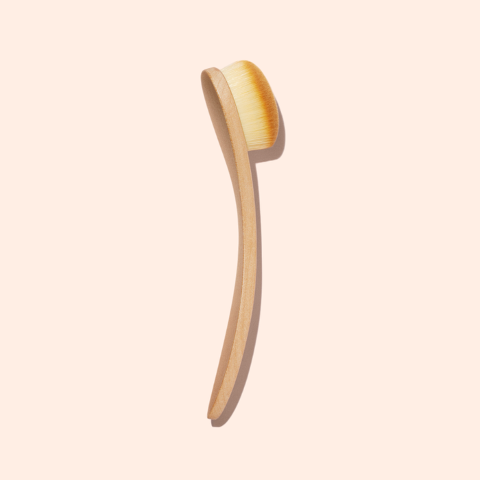 The Manucurist Dust Brush features a wooden body, long curved handle, and soft vegan bristles, shown against a light beige background.