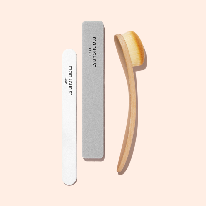 Three manicure tools on a light peach background: a white nail file, gray buffer, and Manucurist Dust Brush with vegan bristles and a wooden handle, each labeled manucurist PARIS.