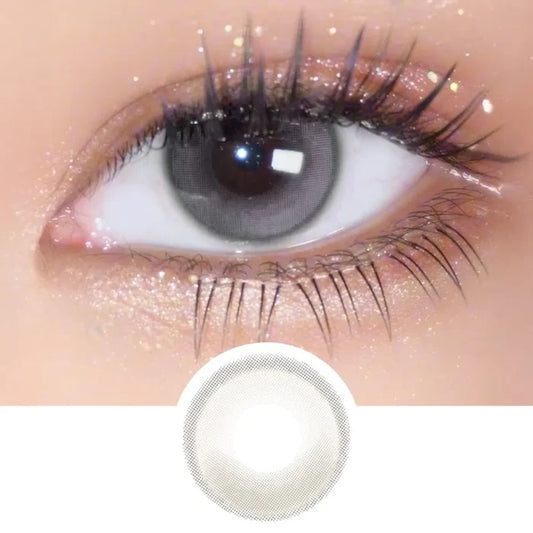 A close-up of an eye with long lashes and shimmery gold eyeshadow wears EyeCandys Amelie Earl Grey circle lenses for a natural look. Below is a circular image showing the design from EyeCandys.