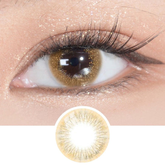 A close-up of an eye with long lashes and shimmery gold makeup wearing EyeCandys Attitude Glitter Brown color contact lenses. Below, a circular image highlights the natural design of these EyeCandys contacts.