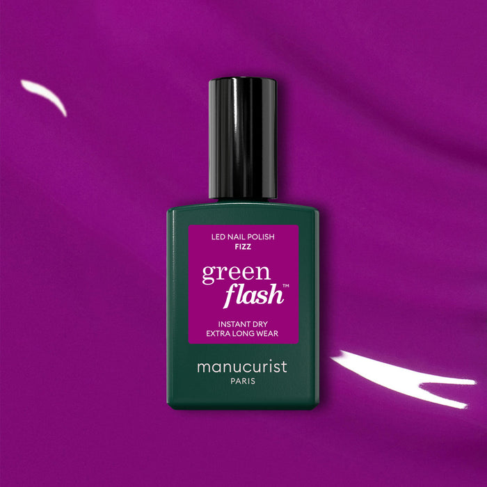 A bottle of Manucurist Fizz Green Flash LED nail polish with magenta undertones stands upright on a vibrant purple background. The label highlights Instant Dry and Extra Long Wear.