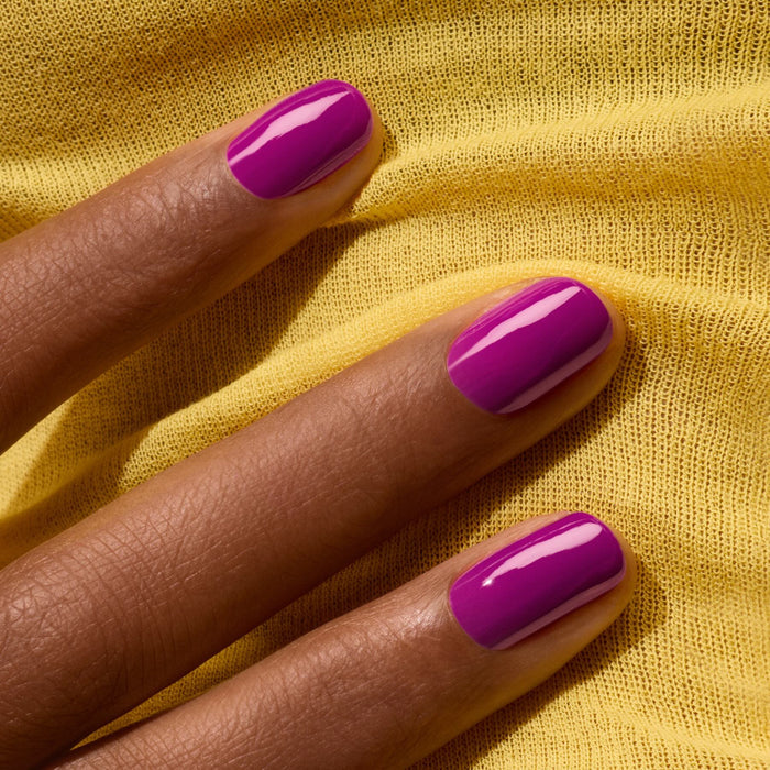 A hand with medium brown skin tone shows neatly manicured nails in Manucurist's Fizz, a semi-matte violet gel polish with magenta undertones, resting on bright yellow textured fabric.