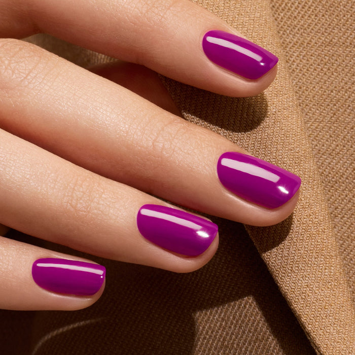 A close-up of a hand with neatly manicured nails painted in Manucurist’s Fizz, a violet polish with magenta undertones, resting on light brown textured fabric.