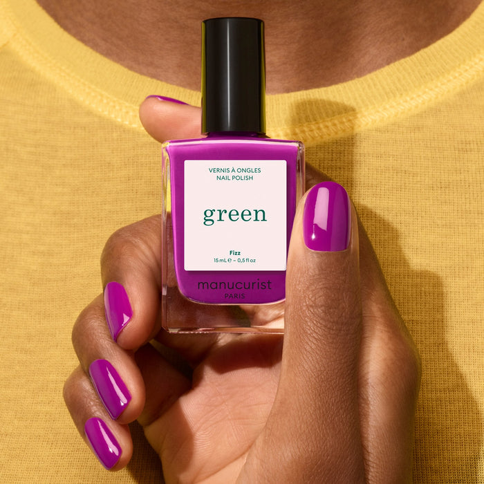 A hand with violet nail polish showing magenta undertones and a semi-matte finish holds a Manucurist Fizz bottle labeled green, set against a yellow shirt background.