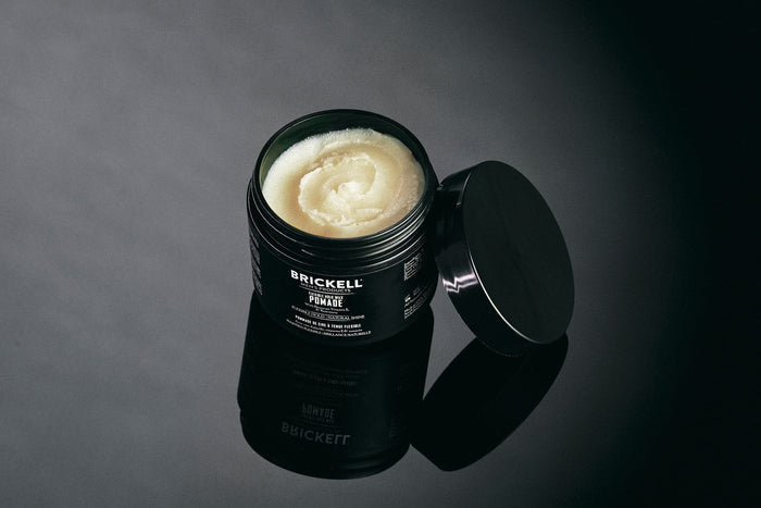 Brickell men's revitalizing hair pomade open container on dark reflective surface with product visible and lid tilted to the side