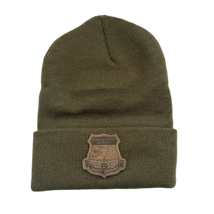 An olive green knit beanie hat with a brown shield-shaped embroidered patch on the cuff, displayed on a white surface.