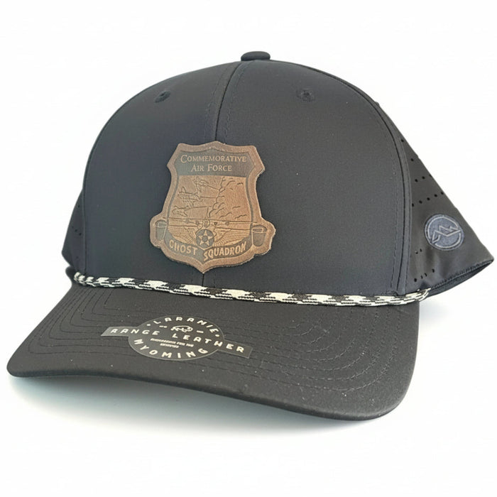 A black baseball cap with a brown shield-shaped patch on the front, a silver braided rope detail across the crown, and a curved brim.