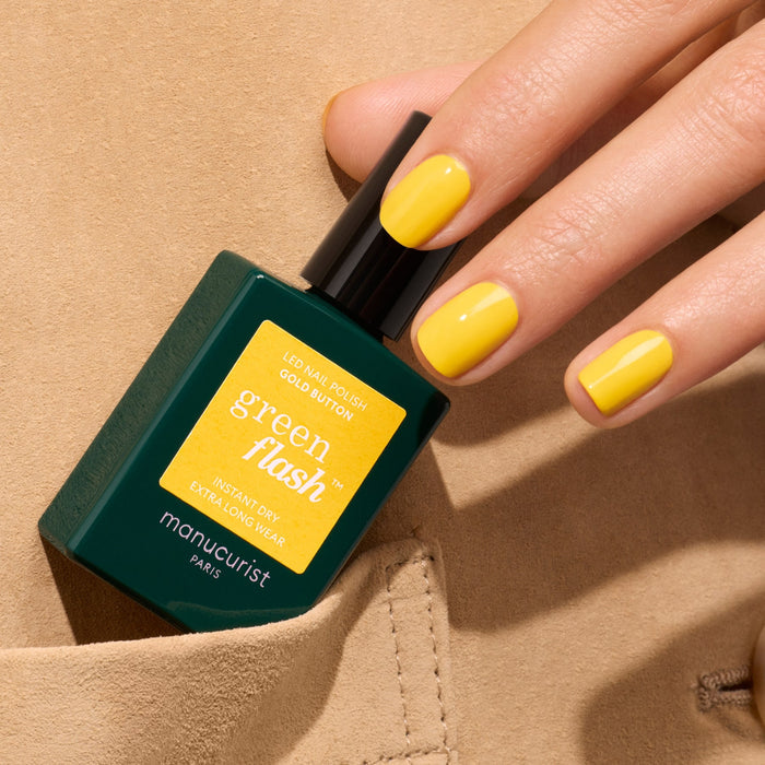 A hand with bright yellow Gold Button nail polish by Manucurist holds a green bottle of Green Flash gel polish, partially tucked into a beige pocket.