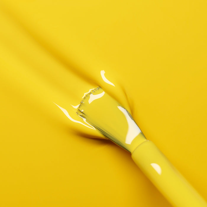 A paintbrush coated in Manucurist’s Gold Button nail polish glides smoothly over a yellow surface, blending perfectly into the background.